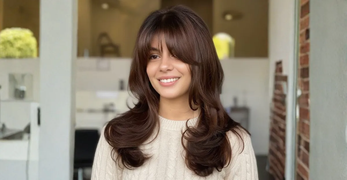 A smiling woman with chocolate brown hair featuring voluminous, flicked-out layers and long, wispy bangs that brush the eyes.